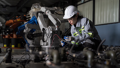 The image shows a woman wearing a hard hat while working on a machine. She appears to be a technician or a blue-collar worker. The setting is indoors.